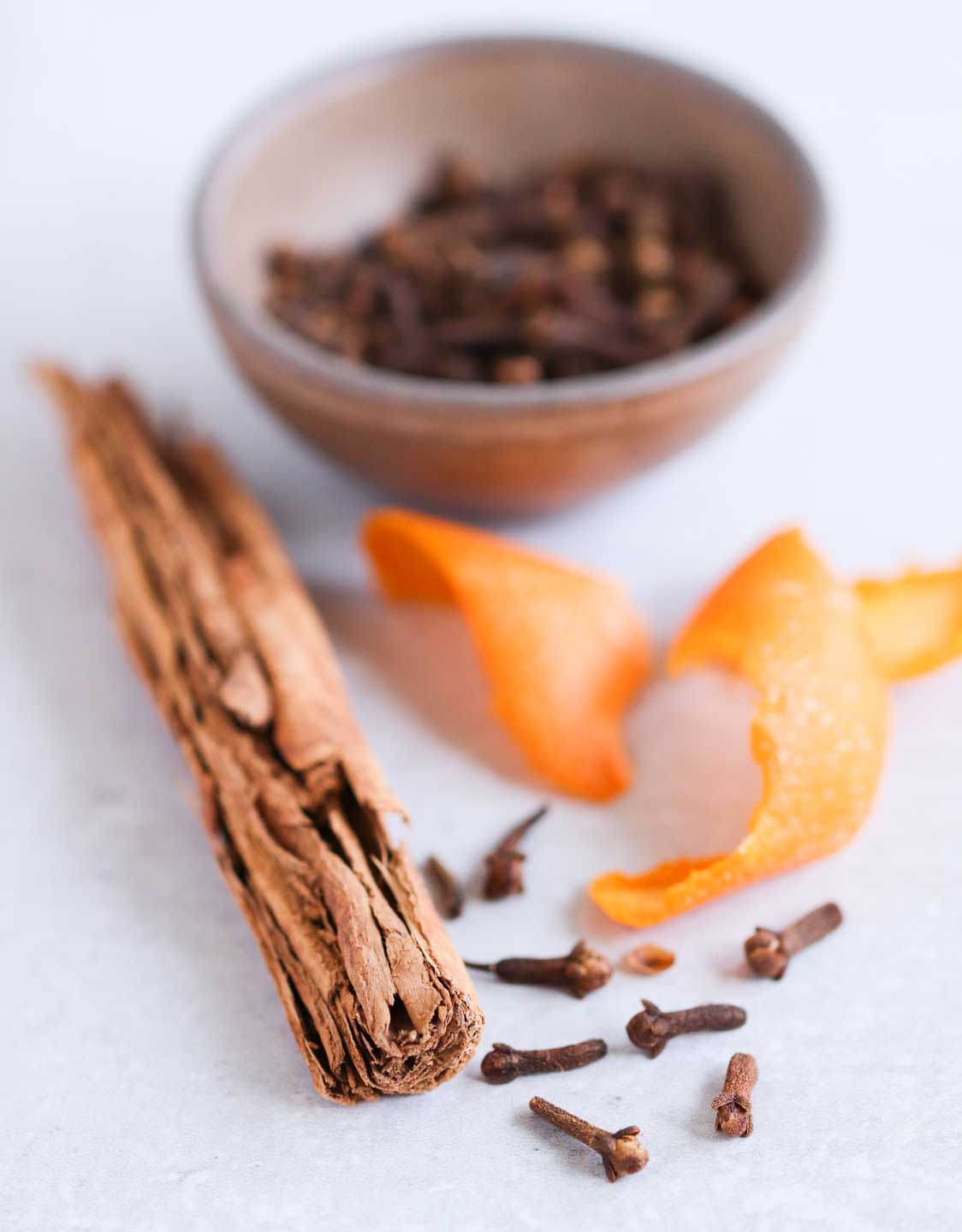 Cinnamon bark, whole cloves and orange peel in the foreground with a bowl of cloves blurred out.