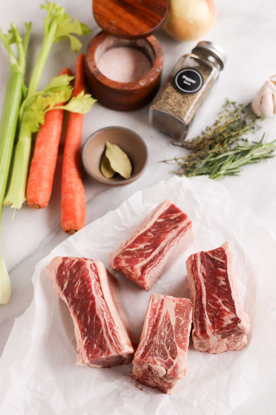 overhead image of uncooked short ribs with vegetables, herbs and spices in the background. 